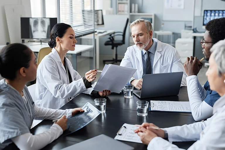female clinician showing medical documents to experienced male doctor sitting by table in front of group of intercultural colleagues